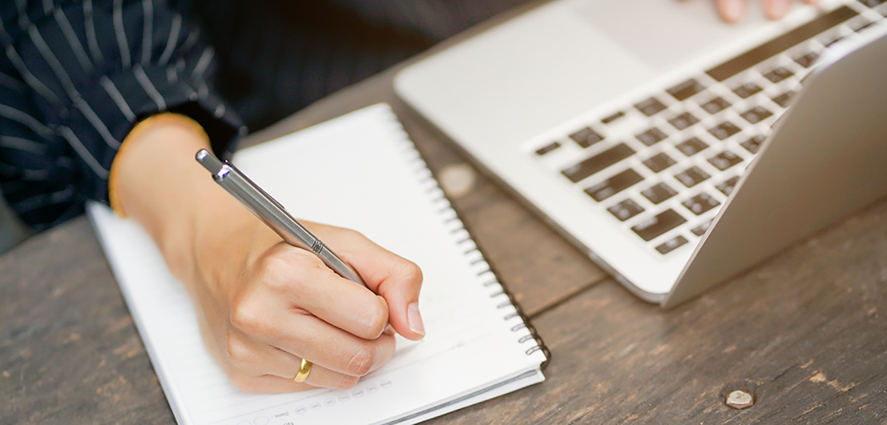 Woman writing in her notebook with laptop open