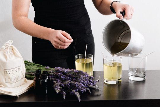 photo of a person in a black dress pouring a soy candle into a glass vessel. lavender is on the table next to the candle along with a linen pouch.