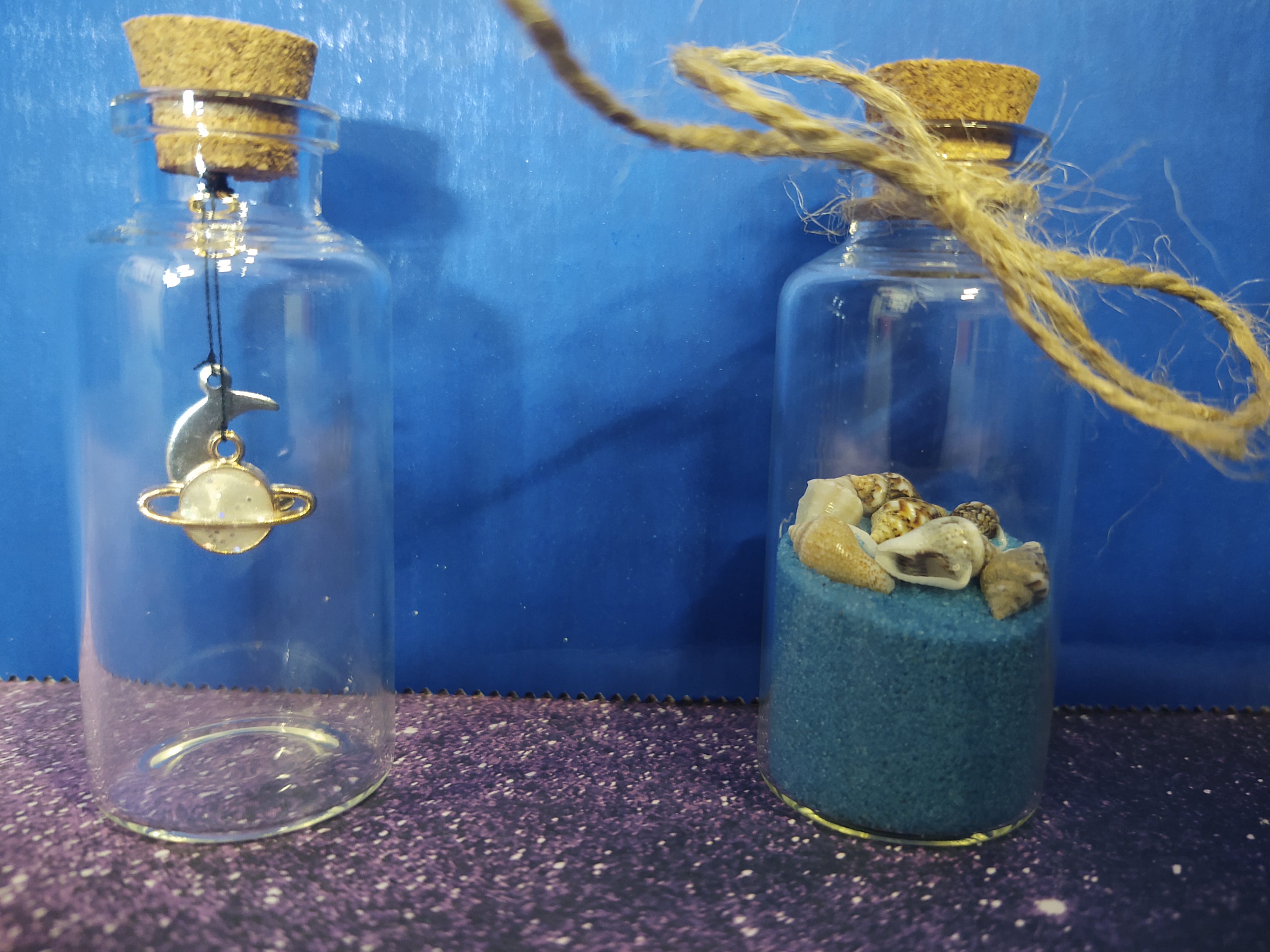 Two bottles, one with a moon and planet charm in it, one with blue sand and seashells with twine tied on top of the bottle.