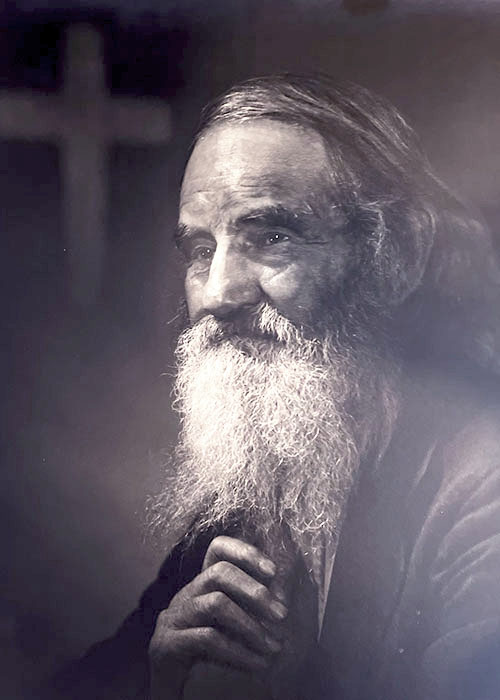 Photographic portrait of an unknown older man with a long white beard and the figure of a cross in the background.