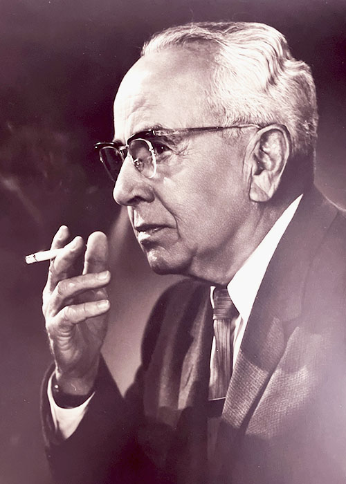 Photographic portrait of an older white man holding a smoking cigarette,