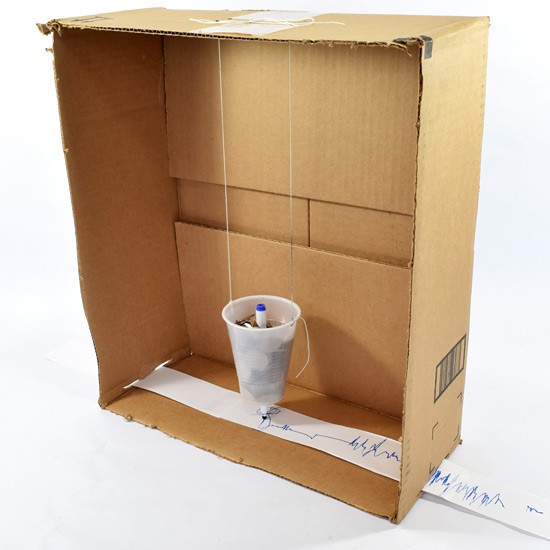 model seismograph made of a cardboard box on its side with cup hanging inside it with strip of paper running underneath