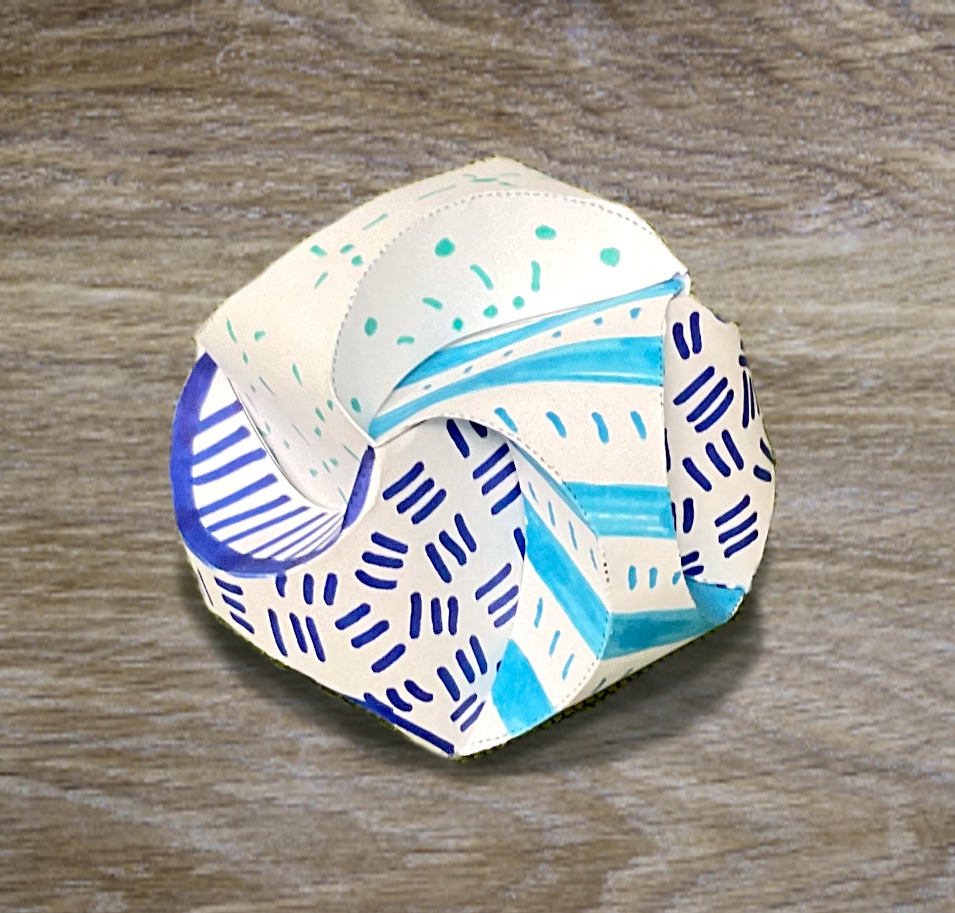 a colorful geometric paper orb with multiple sides that looks like it's swirling. Each face has a different pattern and color: purple hatch marks, green dots and lines, light blue stripes and lines