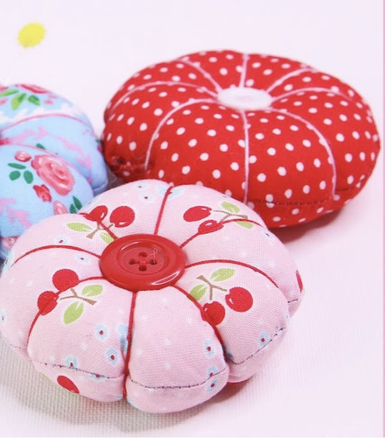 Pincushions in three different styles and colors, a pink pincushion with a red button and cherries; a red pincushion with small white polka dots and a white button, a blue pattern with floral designs