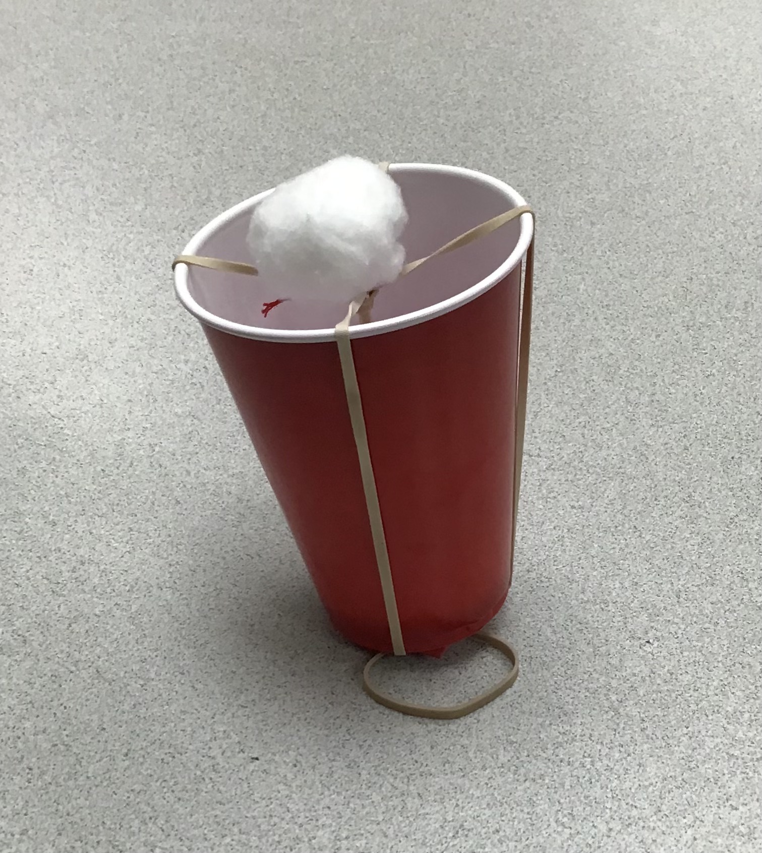 a red plastic cup with 2 rubber bands over the top and more going through the inside to make a launcher, and a cotton ball on top