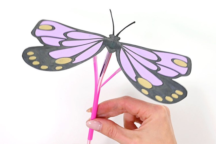 A hand holding a purple paper butterfly with straws underneath to make it flap its wings