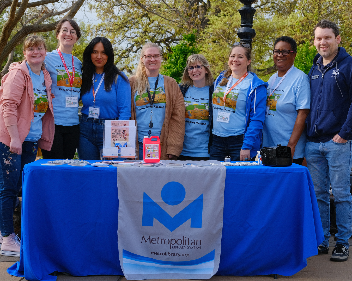Group of 8 people standing behind a table with a blue tablecloth and Metro Library logo.