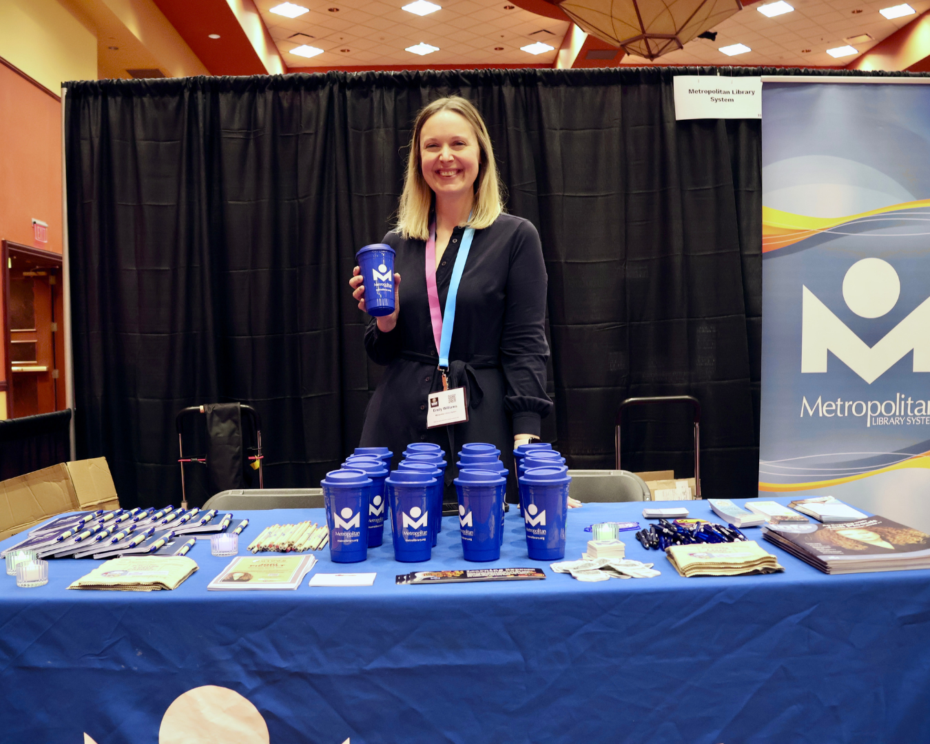 One woman holding a travel coffee mug at a table with the Metro Library logo and various Metro Library-branded items.
