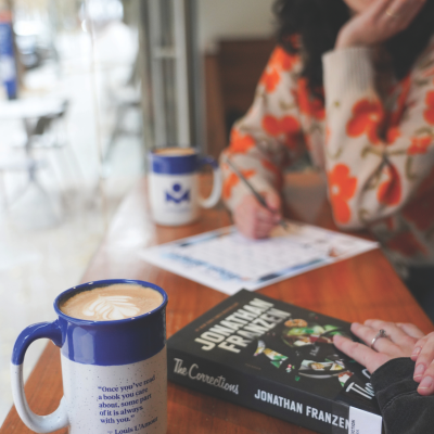 Coffee mug and book on table and a person holding a pen near a piece of paper.