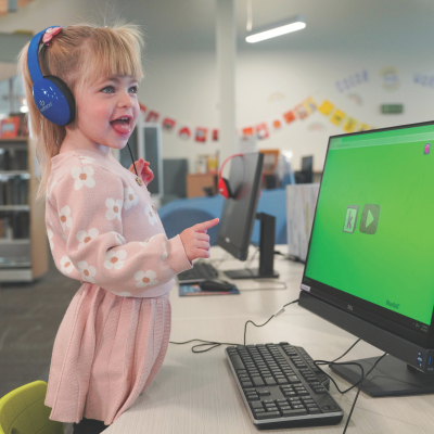 Young girl in a pink sweater and skirt with blue headphones at a computer.