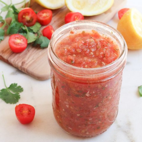 A jar containing red salsa next to a cutting board with tomatoes, cilantro, and lemons on a cutting board