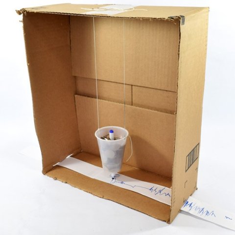 model seismograph made of a cardboard box on its side with cup hanging inside it with strip of paper running underneath