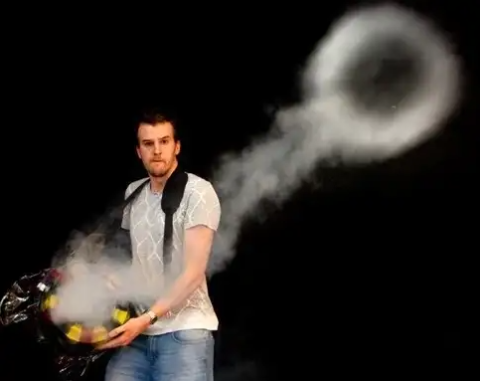 a man holding a large bucket-turned-cannon shooting a smoke ring into the air