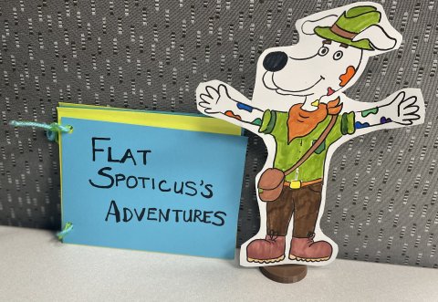 a photo showing a flat cut-out of Spoticus the dog in adventure clothes with a blue and green paper notebook that say's Flat Spoticus's Adventures on it
