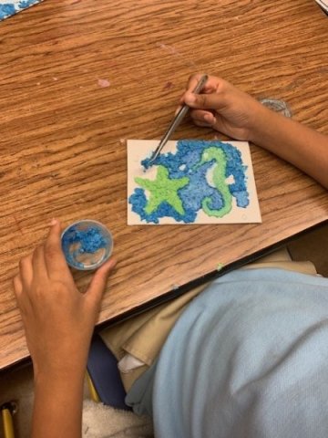 The image depicts a child engaged in an art project at a wooden table. The child is using tweezers to place small blue and green pieces of material onto a piece of white paper. The design on the paper includes a star and a seahorse. Next to the child's left hand, there is a small container holding more blue material.