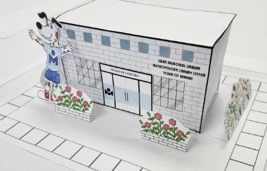 A tiny Harrah Library made out of paper with colorful flowers and Spoticus standing outside.