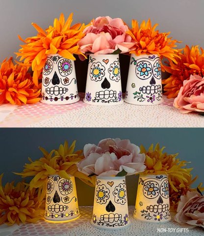 Pictured are three white cups decorated to look like sugar skulls with a battery-powered tea light under them to make them glow. The cups have orange fake flowers on top.