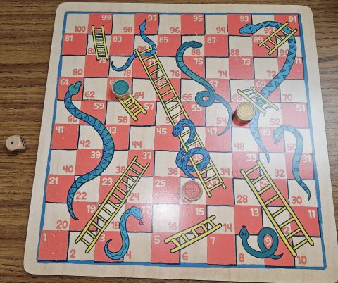 Board game called snakes and ladders with pieces on numbered tiles and a die on the table.