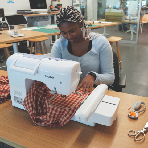 Woman sitting at an embroidery machine working on a project with red and white checkered fabric.