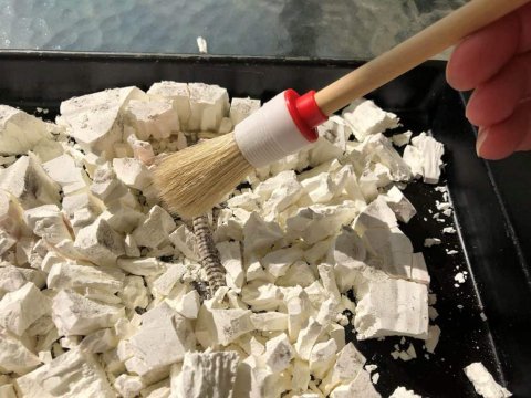 Black tray holding a dinosaur dig that has been broken apart. The guest is holding a small paintbrush and brushing the dirt off the bones. 