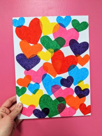 hand holding a small white canvas decorated with multi colored hearts made out of tissue paper against a pink backdrop