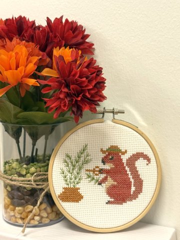 cross stitch of a hat-wearing red squirrel harvesting rosemary from a golden pot.
