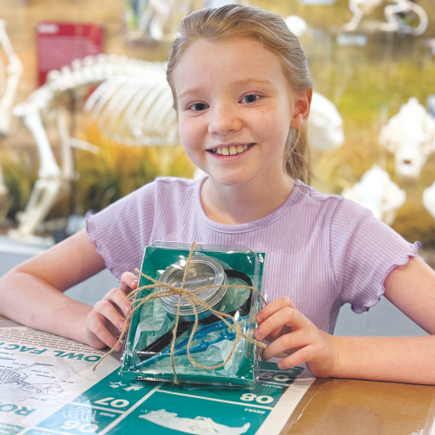 Child holding a green science kit tied with twine, sitting at a table with other materials and a skeleton in the background.
