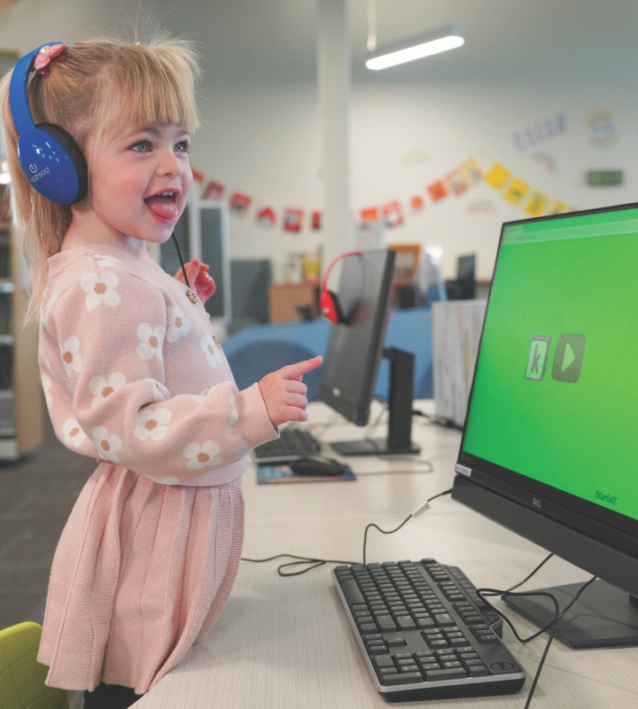 Young girl in a pink sweater and skirt with blue headphones at a computer.